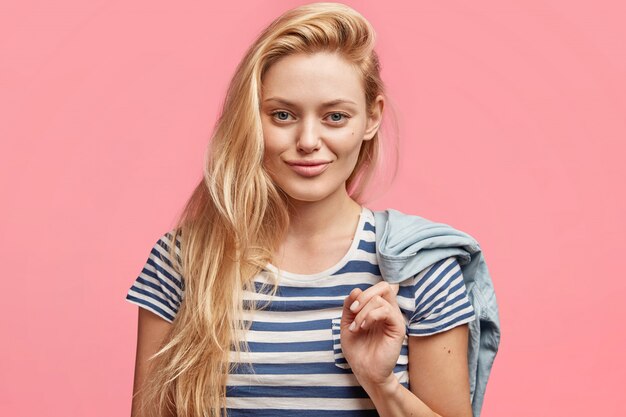 Foto sincera de una hermosa joven guapa con maquillaje, vestida con una camiseta de marinero, sostiene una chaqueta de mezclilla, se ve con expresión alegre y segura