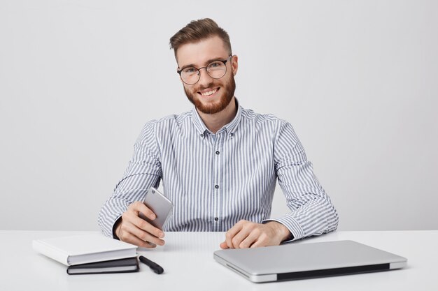 Foto sincera de hombre exitoso con barba espesa, utiliza tecnología moderna para el trabajo