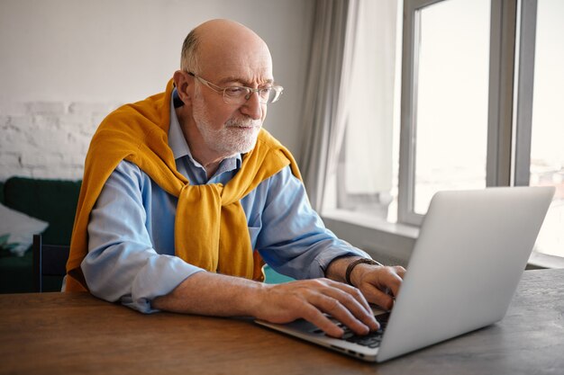 Foto sincera de moda elegante hombre maduro de sesenta años con barba gris y cabeza calva con mirada enfocada, usando una computadora portátil genérica wifi, tecleando rápido. Concepto de personas, edad y gadgets.