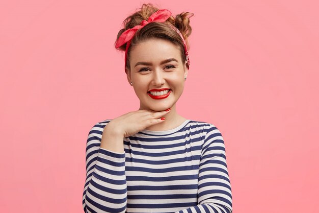 Foto sincera de una mujer de aspecto agradable con maquillaje, expresión positiva, que mantiene la mano debajo de la barbilla, viste un suéter a rayas y una elegante diadema