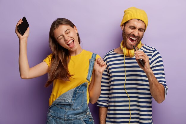 Foto sincera de novios llenos de alegría y energía que escuchan música a través del teléfono celular, bailan y cantan en voz alta, expresan emociones positivas, se paran uno al lado del otro, levantan los brazos y se mueven activamente