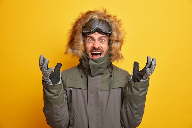 Foto de snowboarder emocionalmente irritado levanta la mano y grita en voz alta expresa emociones negativas usa chaqueta de invierno con gafas de esquí, guantes, gestos mantiene activamente la boca abierta.