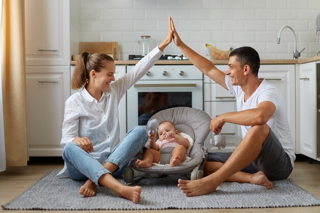 Foto de tamaño completo de positivo, tres personas, mamá, papá, niño pequeño, niña o niño en gorila, los padres hacen las manos en el techo, disfrutando de sentarse en el piso en la cocina ligera, en el interior.