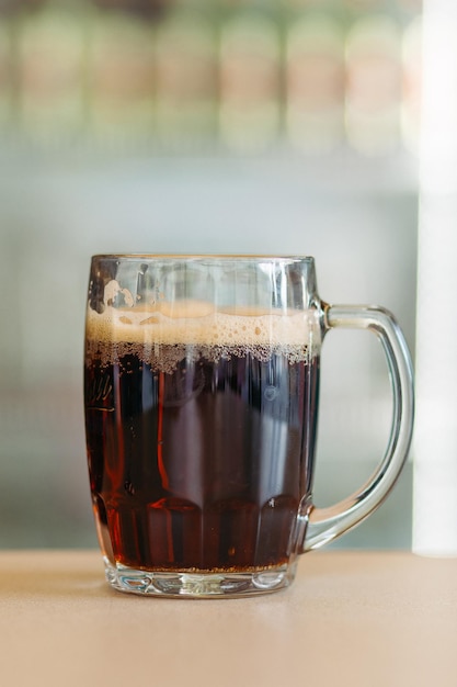 Foto de una taza de cerveza de vidrio de pie sobre una superficie de madera lisa Bebida fría y oscura de verano para el calor del día Fresco con espuma alta y gruesa Fondo borroso claro Concepto de tiro de bebidas alcohólicas ligeras