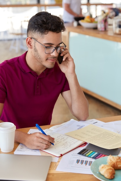 Foto gratuita foto de un trabajador concentrado ocupado con el papeleo, sostiene un bolígrafo, tiene un corte de pelo moderno, vestido con una camiseta informal, se comunica por teléfono celular, disfruta de un delicioso croissant con café. trabajar en la cafeteria