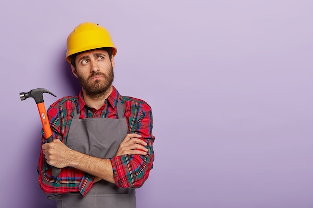Foto de trabajador manual cansado pensativo sostiene un martillo, mantiene los brazos cruzados sobre el pecho, piensa en qué reparar