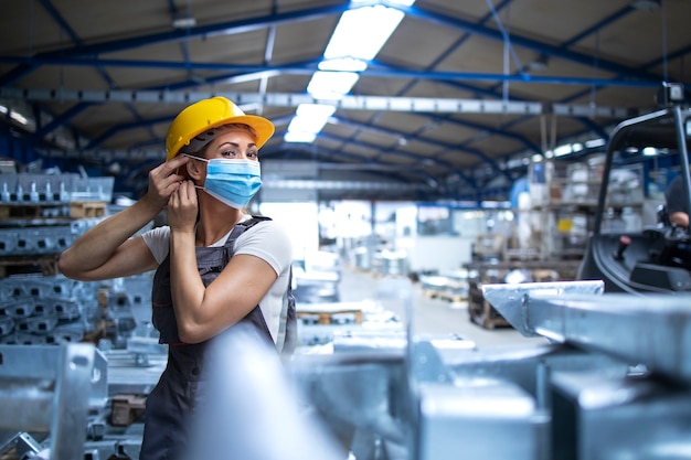 Foto de trabajadora de fábrica en uniforme y casco poniéndose mascarilla en planta de producción industrial