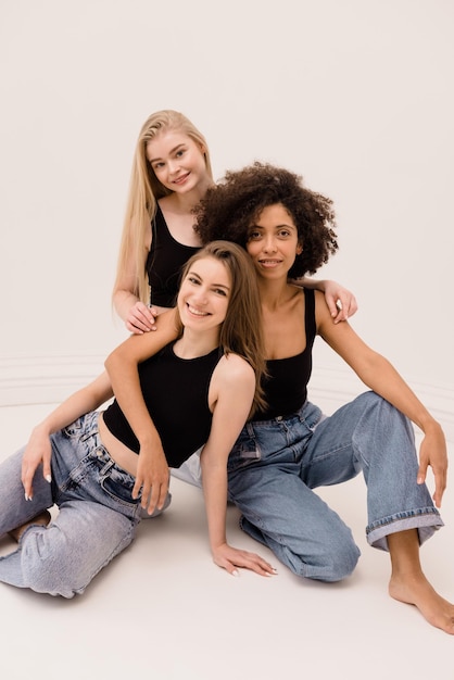 Foto de tres mujeres interraciales jóvenes de cabello claro y oscuro en top negro y jeans aislado sobre fondo blanco.