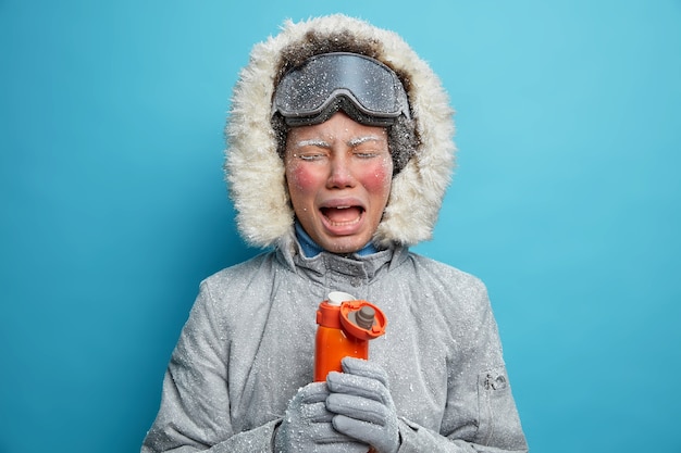 Foto de turista llorando molesta que pasa las vacaciones de invierno activamente siente mucho frío después de ir a esquiar en una tormenta de nieve o ventisca bebe té o café caliente de un termo usa una chaqueta gris con capucha de piel