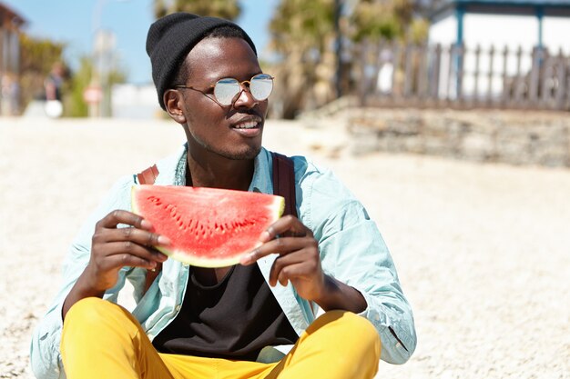 Foto de verano al aire libre del apuesto hombre de piel oscura con ropa de moda y gafas relajantes en la playa durante el día, sentado en una piedra con una rodaja de sandía jugosa, disfrutando de fruta madura y clima cálido