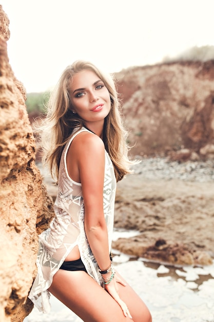 Foto vertical de atractiva chica rubia con cabello largo posando para la cámara sobre fondo de rocas. Ella esta sonriendo.