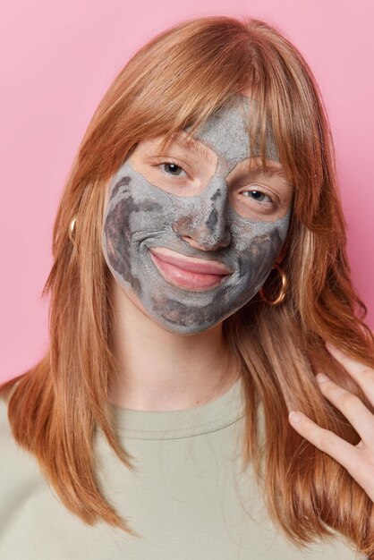 Una foto vertical de una bonita adolescente europea con el pelo de jengibre aplica una máscara de belleza de arcilla para el tratamiento de la piel, limpieza y sonrisas hidratantes, usa aretes y poses de camisetas en el interior