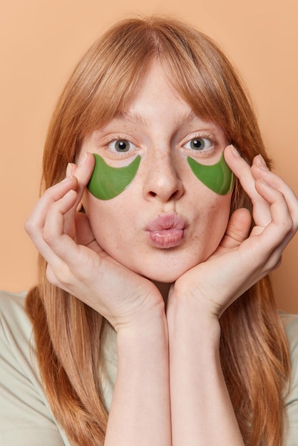 La foto vertical de una chica con pecas de jengibre aplica parches de colágeno verde debajo de los ojos mantiene los labios doblados mira directamente a la cámara se somete a procedimientos de belleza aislados sobre un fondo beige Tratamiento de la piel