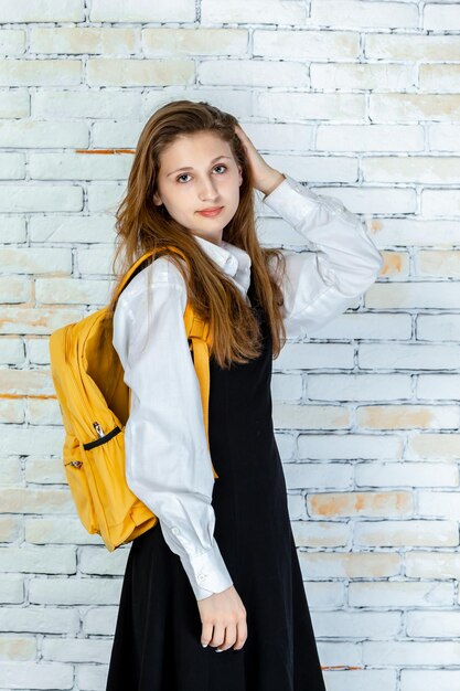 Foto vertical de una estudiante adorable llevándose la mano al cabello y mirando a la cámara Foto de alta calidad