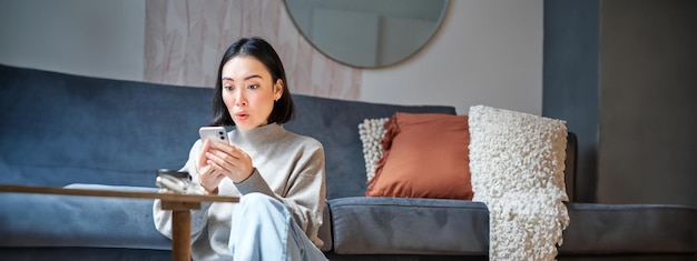 Foto gratuita una foto vertical de una joven asiática parece sorprendida porque su teléfono móvil lee noticias increíbles