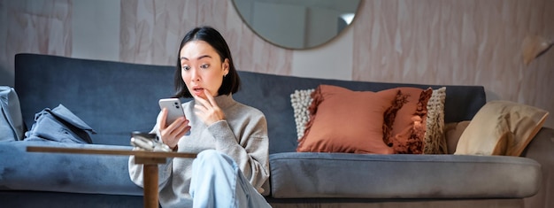 Foto gratuita una foto vertical de una joven asiática parece sorprendida porque su teléfono móvil lee noticias increíbles