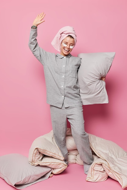 La foto vertical de una joven feliz se divierte despierta después de un largo sueño se siente pacífica y serena usa máscara para dormir y la ropa de dormir sostiene soportes de almohadas suaves contra un fondo rosa lleno de energía