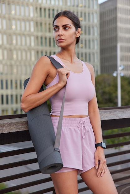 Foto gratuita una foto vertical de una mujer deportiva pensativa vestida con chándal lleva una colchoneta de fitness concentrada en poses a distancia al aire libre contra un fondo urbano borroso. mujer atlética va a tener pilates