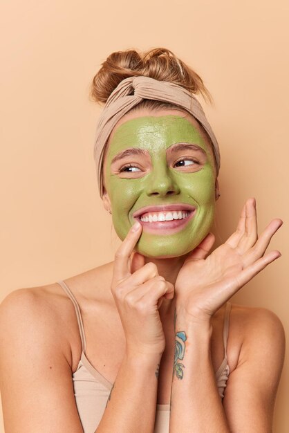 La foto vertical de una mujer europea feliz sonríe con dientes y aplica una máscara de belleza natural verde para el tratamiento de la piel usa una camiseta informal y una banda para la cabeza aislada sobre fondo marrón Concepto de cuidado facial