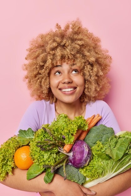 Una foto vertical de una mujer feliz y curiosa sonríe con dientes y mira por encima de los avisos de que algo atractivo abraza un gran montón de vegetales verdes frescos que mantienen una dieta saludable aislada sobre un fondo rosado