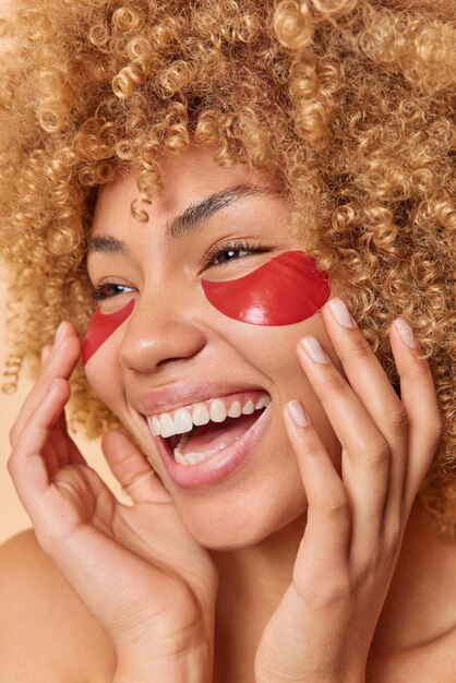 La foto vertical de una mujer feliz con el pelo rubio rizado aplica parches de colágeno rojo para el tratamiento de la piel toca la cara suavemente se siente alegre elimina las arrugas sonríe ampliamente. Fotografía de cerca. tiempo de belleza