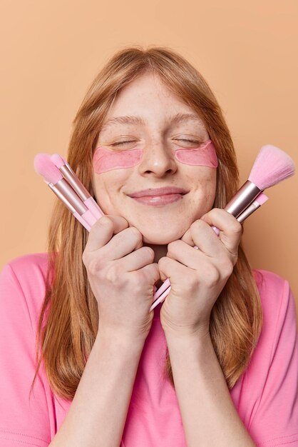 La foto vertical de una mujer guapa y complacida con el pelo largo y rojo mantiene los ojos cerrados aplica parches de hidrogel sostiene cepillos cosméticos se somete a procedimientos de belleza para el cuidado de la piel aislados sobre una pared beige