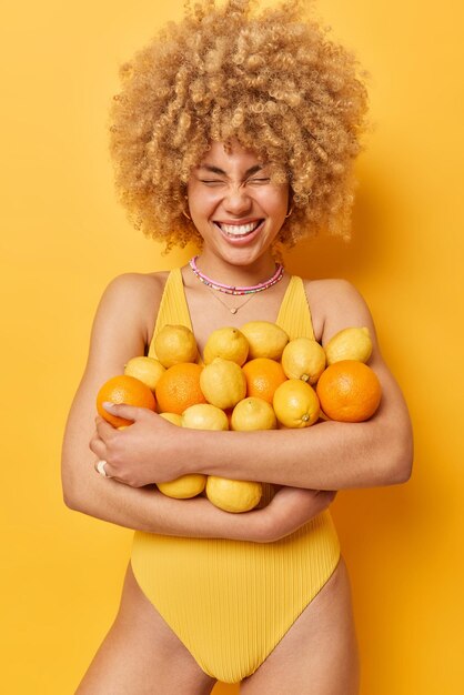 Una foto vertical de una mujer de pelo rizado positiva abraza un gran montón de jugosas frutas tropicales cítricas sostiene naranjas y limones vestidos con traje de baño sonríe positivamente aislado sobre un fondo amarillo vívido