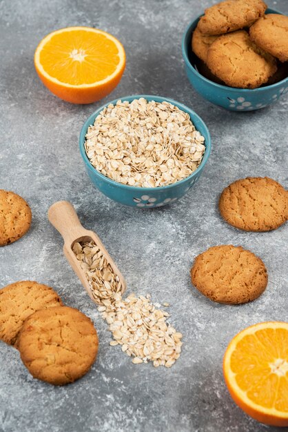 Foto vertical de pila de galleta y avena en un bol y naranja medio cortado sobre una superficie gris.