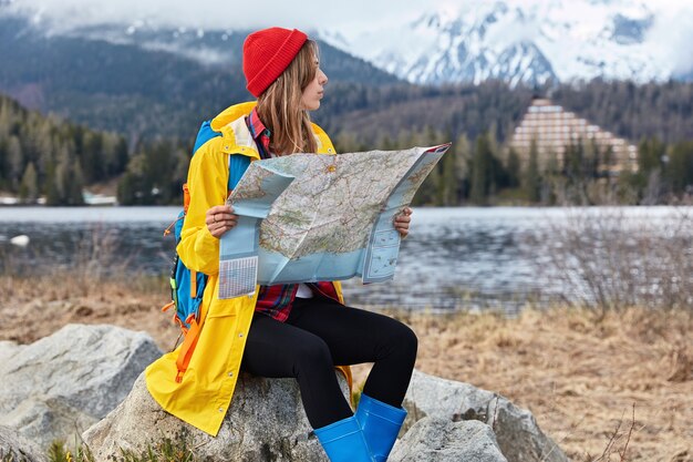 Foto de viajera seria con mochila explora un nuevo destino, lee el mapa mientras se sienta en una piedra, busca algún lugar