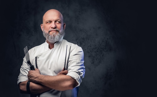 Una foto de un viejo chef profesional vestido con uniforme sosteniendo una espátula de cocina y pinzas.