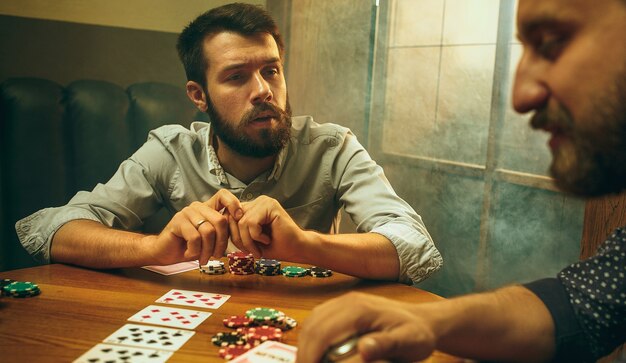 Foto de vista lateral de amigos masculinos y femeninos sentados en la mesa de madera. Hombres y mujeres jugando al juego de cartas. Manos con primer plano de alcohol.