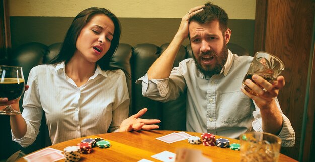 Foto de vista lateral de amigos masculinos y femeninos sentados en la mesa de madera. Hombres y mujeres jugando al juego de cartas. Manos con primer plano de alcohol.