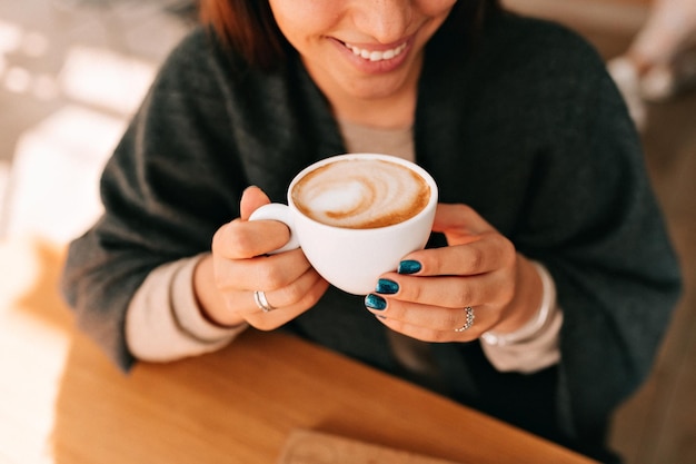 La foto de la vista superior de una dama adorable sonriente con una maravillosa sonrisa manicura oscura está tomando café y disfrutando descansando en la cafetería