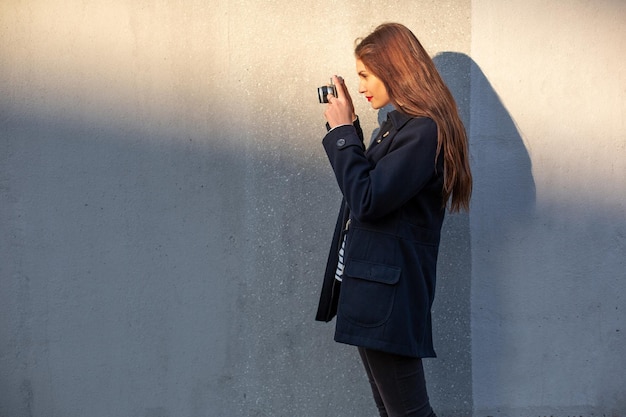 Fotógrafa sonriente con chaqueta parada frente a la pared lista para hacer una nueva foto. Adorable mujer morena joven en traje de moda posando sobre fondo de pared de hormigón con cámara