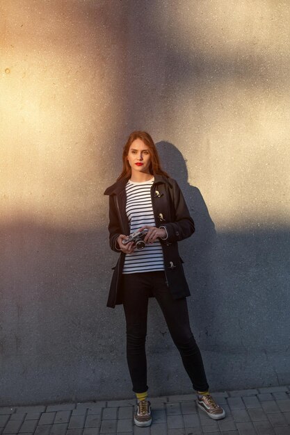 Fotógrafa sonriente con chaqueta parada frente a la pared lista para hacer una nueva foto. Adorable mujer morena joven en traje de moda posando sobre fondo de pared de hormigón con cámara