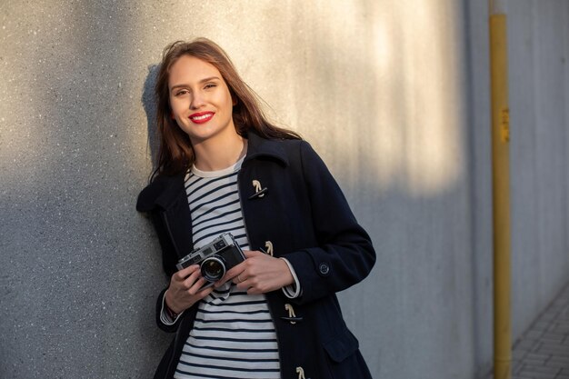 Fotógrafa sonriente con chaqueta parada frente a la pared lista para hacer una nueva foto. Adorable mujer morena joven en traje de moda posando sobre fondo de pared de hormigón con cámara