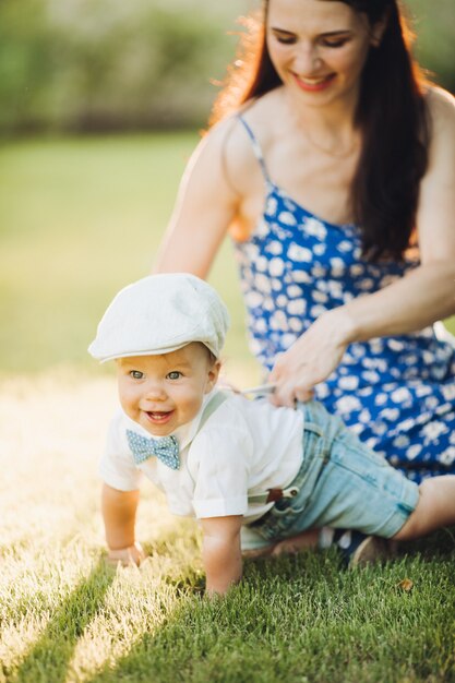 Fotografía de archivo retrato de un bebé adorable y dulce con elegante sombrero, lazo y pantalones cortos de mezclilla con camisa arrastrándose sobre el césped mientras su madre lo sostiene y sonríe. Actividad de verano juntos al aire libre.