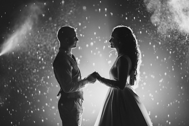 Fotografía en blanco y negro de la alegre novia y el novio tomados de la mano y sonriendo el uno al otro contra los fuegos artificiales brillantes