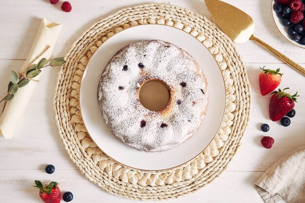 Fotografía cenital de un anillo de pastel con frutas y polvo sobre una mesa blanca con fondo blanco.