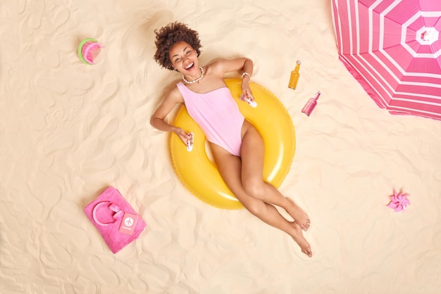 Fotografía cenital de una feliz mujer de cabello rizado en traje de baño posa en un anillo de natación inflado amarillo pasa tiempo libre en la playa yace al sol rodeada de juguetes de arena botella de bebidas energéticas auriculares en una toalla