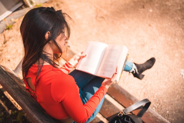 Fotografía cenital de una joven morena caucásica con una blusa roja leyendo un libro en un banco del parque