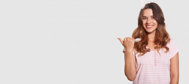 La fotografía interior de una mujer relajada y de aspecto agradable anuncia algo, apunta con el pulgar al espacio en blanco, muestra espacio libre para su contenido promocional, tiene una sonrisa encantadora y atractiva en la cara