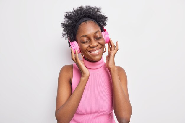 Fotografía en interiores de una encantadora mujer de piel oscura que cierra los ojos por placer, escucha su música favorita, mantiene las manos en los auriculares estéreo de color rosa