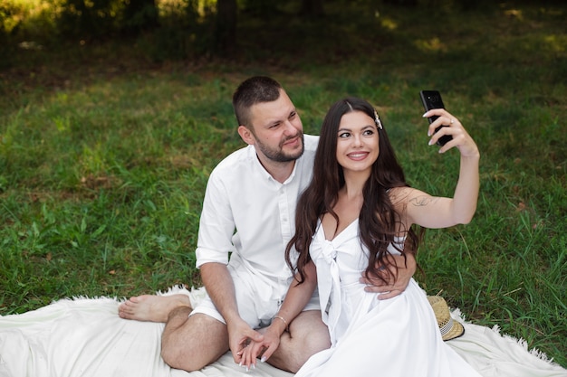 Fotografía de stock de una hermosa pareja en ropa blanca sentada en una manta de picnic. bonita novia con cabello largo castaño en vestido blanco sosteniendo teléfono móvil y tomando selfie.