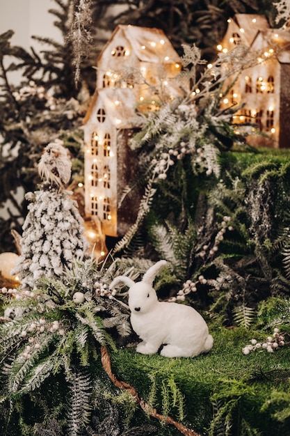 Fotografía de Stock de hermosas decoraciones navideñas con ramas de abeto y conejo blanco de juguete. Casas artesanales iluminadas.