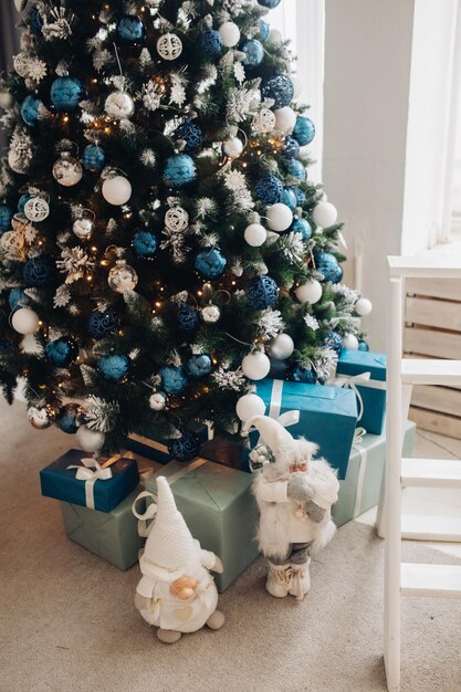 Fotografía de Stock de hermoso árbol de Navidad decorado con bolas azules y plateadas y blancas y regalos de Navidad envueltos bajo el árbol. Dos figuras de Papá Noel debajo de un árbol.