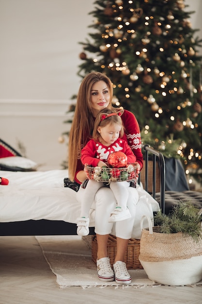Fotografía de Stock de madre amorosa en vestido verde dando a su pequeña hija en pijama vestido un regalo de Navidad. Están al lado de un árbol de Navidad bellamente decorado bajo la nieve.