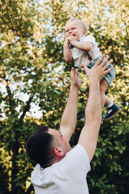 Fotografía de Stock de un padre amoroso lanzando a su hijo en el aire contra el gran árbol verde en la luz del sol. Niño feliz con su padre. Concepto de familia.