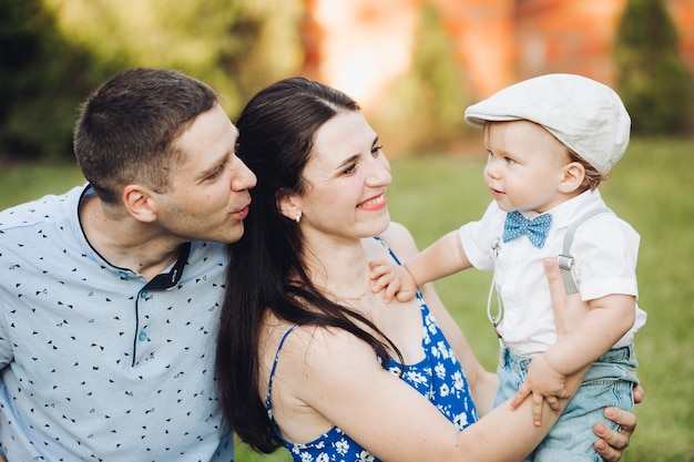 Fotografía de Stock de papá y mamá con hijo en el parque. Hermosa madre sonriente sosteniendo a su hijo en brazos, padre sonriéndole detrás de la mamá. Niño con sombrero y moño. Concepto de familia feliz.