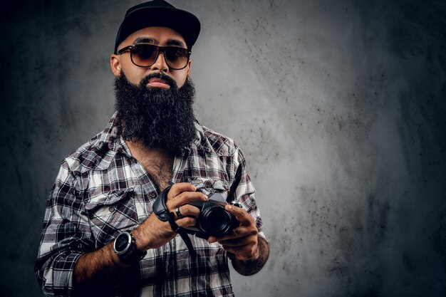 Un fotógrafo aficionado hipster barbudo con tatuajes en los brazos, vestido con una camisa de lana sostiene una cámara réflex digital compacta sobre fondo gris.